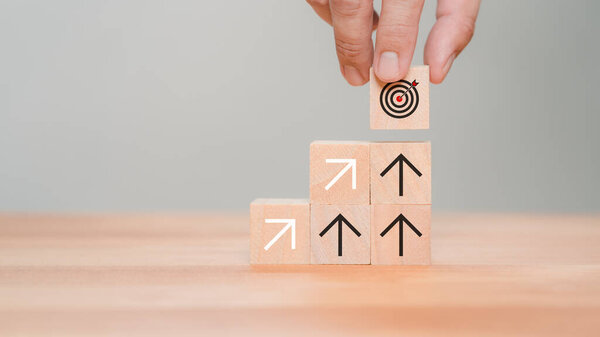 Male hand put a wooden cube block with the goal icon on the top of stacking wooden cubes standing with arrow up icon. Business concept of goal achievement, financial growth, target and investment.