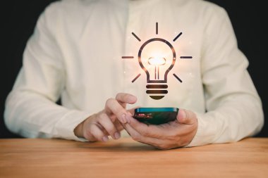 Parlayan bir ampul ikonuyla akıllı telefon tutan bir kişinin yakın çekimi, yeniliği, yaratıcılığı ve yeni fikirleri sembolize ediyor. Bu dijital kaplama ilham ve teknolojik gelişmeyi öneriyor..