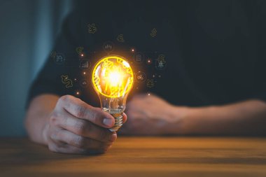 Bir kişi para birimi sembolleri ve iş simgeleriyle çevrili, yenilikleri, finansal fikirleri ve iş ve teknolojide yaratıcı çözümleri sembolize eden parlak bir ampul tutuyor..