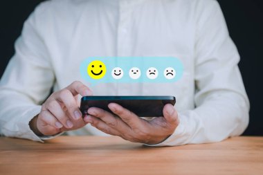Akıllı telefon tutan kişi, çevrimiçi geri bildirim sisteminde müşteri memnuniyetini, gözden geçirmeyi ve pozitif kullanıcı deneyimini sembolize eden, beş emoji seçeneğine sahip bir geri bildirim skalasından mutlu yüz simgesi seçer..