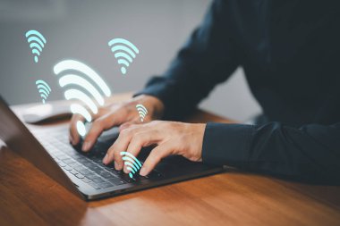 Kablosuz internet bağlantısını, çevrimiçi iletişimi, uzaktan erişimi, uzaktan erişimi ve dijital çalışma alanındaki ağ bağlantısını sembolize eden yüzen Wi-Fi simgelerine sahip dizüstü bilgisayar kullanan bir iş adamı.