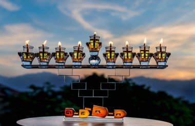 Kristal menorah ve zeytin yağı ile yanan mumlar - Hanuka 'nın sembolü - Musevi Mucize Işık Tatili. Dağların ve gökyüzünün bulanık arkaplanı, İbranice harfler - burada büyük bir mucize gerçekleşiyor.