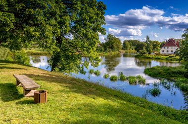 Gölde güzel yansımaları olan eski bir halk parkı, ön planda park bankı ve küçük binalı bir ada, aydınlık bir yaz günü.