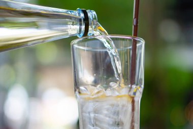 Taze dökülmüş berrak, pırıl pırıl, şeffaf bir bardağa akan sıvı bir içecek bulanık yeşil bir arka plana karşı. Doğada soğuk ve taze bir içeceğin tadını çıkarma kavramı..