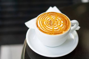 Beyaz seramik bir fincanda, ahşap bir kafe masasında, zarif bir şekilde kıvrılmış latte sanat tasarımı. Köpüklü, kremalı içecek mükemmel bir sabah içkisi ya da öğlen molası..