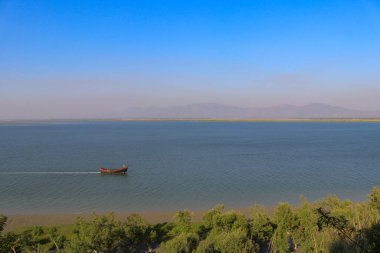 Geleneksel bir balıkçı teknesi, yeşil kıyı şeridi ve açık mavi gökyüzünün altındaki uzak dağların yer aldığı Bangladeş ve Myanmar arasında akan Naf Nehri 'nin sakin bir görüntüsü..