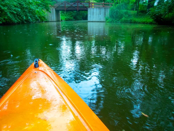 Bright orange kayak travels leisurely down a beautiful tranquil river with bridge in the distance surrounded by lush green vegetation and reflection in water. No people, shot in natural light with