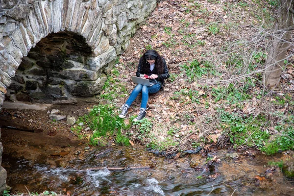 早春的自然场景 一个年轻人带着耳机 一个人坐在流淌的溪流边 一个带着笔记本电脑的石桥 手里拿着一个红色麦克风 自然光 复制空间 — 图库照片