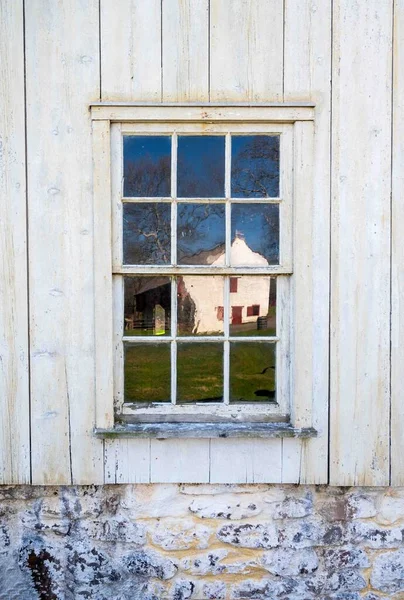 Exterior colonial Pennsylvania farm house window with wavy antique glass reflects a large whitewashed stone barn. Window is in a rustic white wooden exterior wall with whitewashed weathere stone foundation. No people, Copy space.