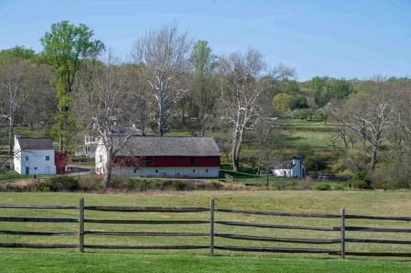 A wooden split rail fence in foreground across green grass pasture with historical colonial american village scene background with agricultural buildings, homes, general store, and orchard landscape and woods beyond. No people, with copy space