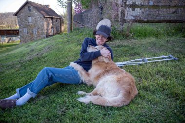 Arka planda, genç bir adam ve sevgili Golden Retriever köpeğiyle birlikte antik taş binalarla dolu çimenli bir tepede uzanan bu sakin kırsal doğa sahnesinin koltuk değnekleri.