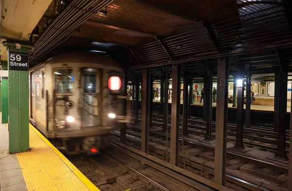59. caddedeki New York metro istasyonunda yeşil sütun ve sarı güvenlik hattı var. Metro vagonu geliyor, hareket bulanıklığıyla önde görünüyor.