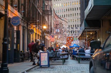 New York, New York, ABD 10-05-2023. Sokakta masalar ve banklar dolu. New York 'ta bir caddedeki Oktoberfest için ışıklar asılmış ve simit süsleri asılmış. Altın saat ışığı, olumlu duygu, yemek saatinde mutlu insanlar. Sadece editör kullanımı.