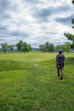 A young man walks toward the Native American Hopewell Culture prehistoric Seip Earthworks burial mound in Ohio. Ancient large long mound. Grass is neatly trimmed with trees and dramatic sky. Copy