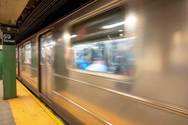 59. caddedeki New York metro istasyonunda yeşil sütun ve sarı güvenlik hattı var. Metro vagonu geliyor ya da kalkıyor hareket bulanık yolcuları pencerelerden görülebilir.