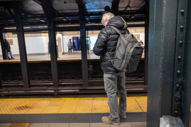 New York, New York, ABD 02-19-2023 Off merkez erkek yolcu kışlık palto ve sırt çantası ile Manhattan metro istasyonunda tren bekliyor, diğer yolcular da görülebilir durumda. Sarı güvenlik hattı ve çelik kirişler. Sadece editör kullanımı