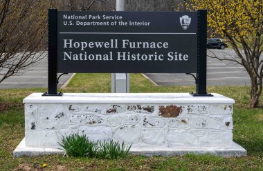 Hopewell Furnace Ulusal Parkı, Pennsylvania, ABD, 03-13-2024. Çimlerle ve çiçek açan çiçeklerle giriş işareti. ABD İçişleri Bakanlığı, Ulusal Park Hizmetleri.
