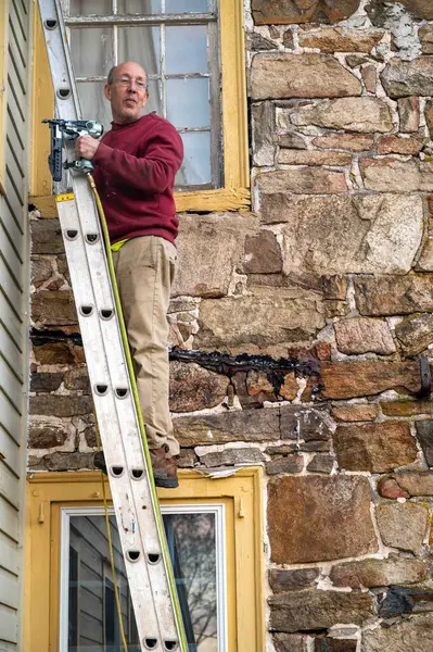 Man high up on a ladder leaning against an old filedstone farmhouse with antique windows. He carries a pneumatic brad gun in his hand