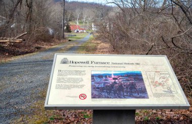 Elverson, Pennsylvania, USA 03-13-2024 Hopewell Furnace National Historic Site informational sign along gravel path into the village. rural landscape background with distant cottages and buildings clipart