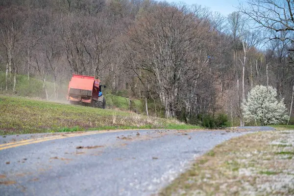 一辆蓝色的农场拖拉机拖着一辆红色的粪肥播种机肥料拖车停在乡间小路旁的绿色草地上 具有分散的前景 复制空间和早春林地背景 — 图库照片