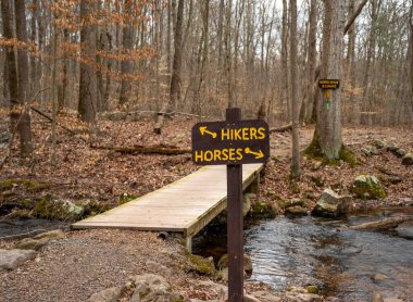 Elverson, Pennsylvania, ABD 03-15-2024 At nalı ve Lenape patika işaretleri Pennsylvania 'daki Fransız Creek Eyalet Parkı' nda akan akarsu ile ahşap bir yaya köprüsünün üzerinden atların ve yürüyüşçülerin yolunu gösteriyor.