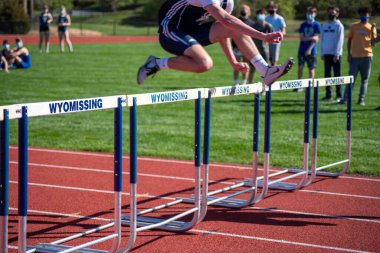 Wyomissing, PA, USA 04-13-2021 Young student athlete leaps over a hurdle on an athletic running track in an outdoor track and field event competition. Motion blur and defocused spectators in clipart