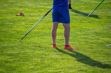 04-20-2021 Mavi üniformalı ve turuncu ayakkabılı genç bir atlet altın saat çimenli bir atletizm sahasında cirit atıyor. Sadece editör kullanımı