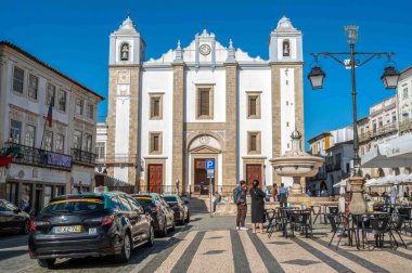 Evora, Portekiz 11-19-2024 Sao Antao Kilisesi 'nin önündeki açık hava restoranlarında toplanan insanlar park halindeki şemsiyeler ve parke taşlı kasaba meydanındaki mağazaların önünde toplanıyor.