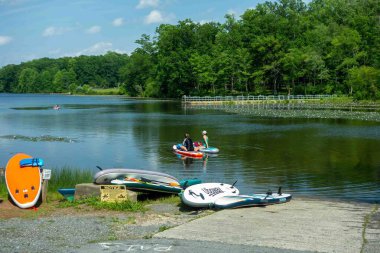 French Creek State Park, Elverson, PA 06-10-2025 Paddle sörf tahtaları ve güvenlik levhaları arka planda iki kayıkla huzurlu bir gölün çimenli sahilinde hafta sonu tatili tatili rahatlama