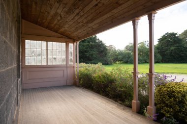 Kereste zemin ve çatısı olan ahşap bir verandanın arka plan dokusu taştan bir kır evinin önünde geniş yeşil bir alana bakıyor. Tasarımınız için alanı kopyalayın. 