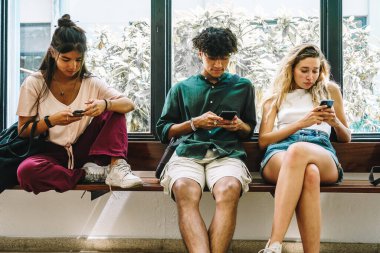 Çeşitli erkek ve kadınlar gündelik giysiler içinde pencerenin yanındaki bankta oturuyor ve üniversite binasının geniş koridorunda ders beklerken cep telefonlarını karıştırıyor.