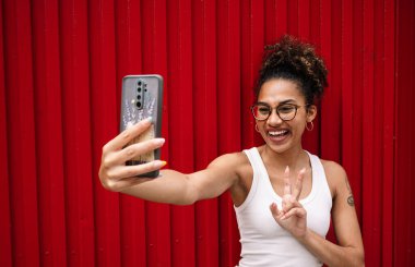 Beyaz bluzlu mutlu Afro-Amerikan kadın ve gözlüklü V işareti ve kırmızı duvara karşı akıllı telefonla selfie çekerken gülümsüyor.