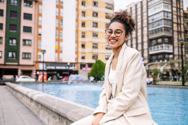 Gündüz vakti şehrin göbeğinde havuz kenarında otururken kameraya gülümseyen şık giysili ve gözlüklü Afro-Amerikan kadın.