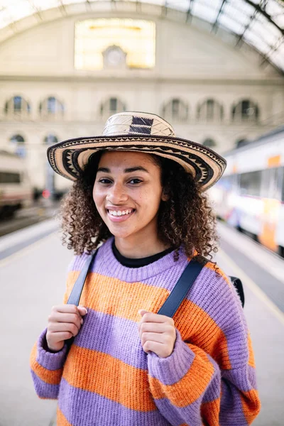 Positive young female in casual outfit and straw hat looking away while standing near train on contemporary railway station