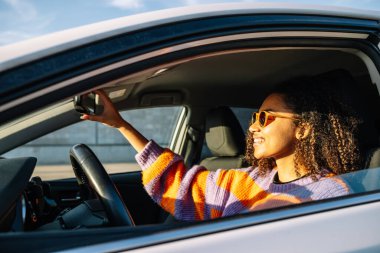 Sıcak kazaklı, Afro saç stili neşeli genç bir kadın. Şoför koltuğunda oturmuş aynayı ayarlıyor ve güneşli bir günde gülümsüyor.