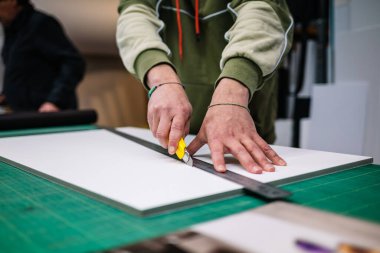 Crop anonymous male worker in casual clothes cutting board with knife at workbench in printing industry