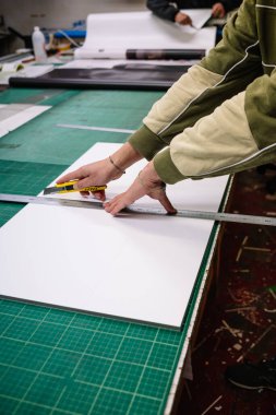 Crop anonymous male worker in casual clothes cutting board with knife at workbench in printing industry