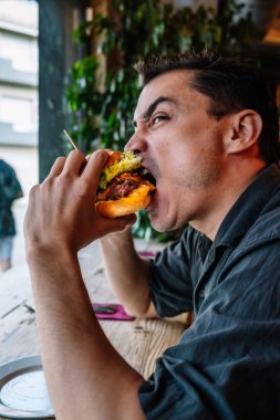 Gündüz vakti restoranda öğle yemeği sırasında pencerenin yanındaki masada oturan aç bir adam lezzetli bir burger yiyor.