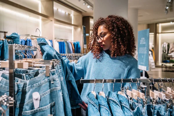 Günlük kıyafetlerle kıvırcık saçlı Afro-Amerikalı genç kadın alışveriş merkezini ziyaret ederken kot pantolon seçiyor.