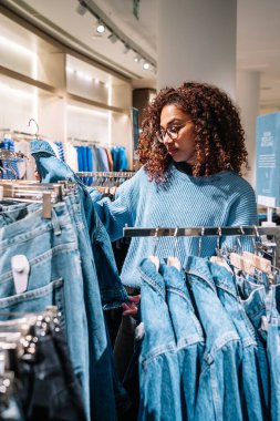 Günlük kıyafetlerle kıvırcık saçlı Afro-Amerikalı genç kadın alışveriş merkezini ziyaret ederken kot pantolon seçiyor.
