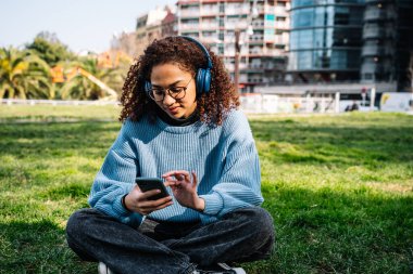 Odaklanmış kıvırcık saçlı, gündelik giysiler ve gözlüklü, kulaklıkla çimenli çimlerde oturan, cep telefonuyla mesajlaşan ve parkta müzik dinleyen bir kadın.