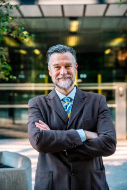 Resmi ceketli ve kravatlı, gri sakallı, kendine güvenen yetişkin bir işadamı kameraya bakıyor ve kollarını kavuşturarak şehir caddesinde dikilirken gülümsüyor.