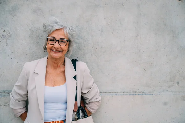 Smiling elderly female wearing eyeglasses and beige jacket standing with hands behind back leaning on concrete wall looking at camera