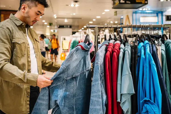 stock image Side view of crop Asian male customer selecting denim jacket hanging on rack while shopping in modern mall