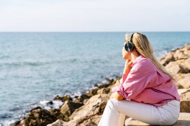 Denize yakın oturan ve kayalık sahilde şarkı söylerken kulaklıkla müzik dinleyen genç bir kadının yan görüntüsü.