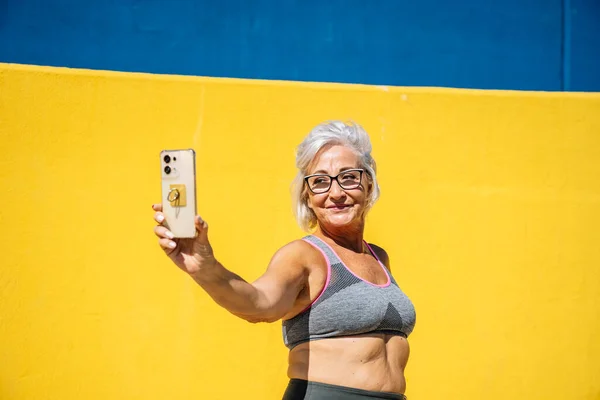 Happy and proud senior woman in sports clothes taking a selfie in a yellow background