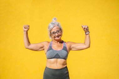 Yaşlı, mutlu, sportif bir kadın sarı arka planda kaslarını ve gücünü gösteriyor.