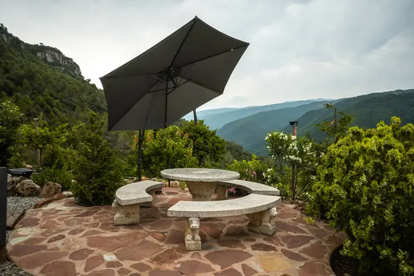 Outdoor stone patio featuring a round table with benches and a large umbrella, set against a lush mountain backdrop.