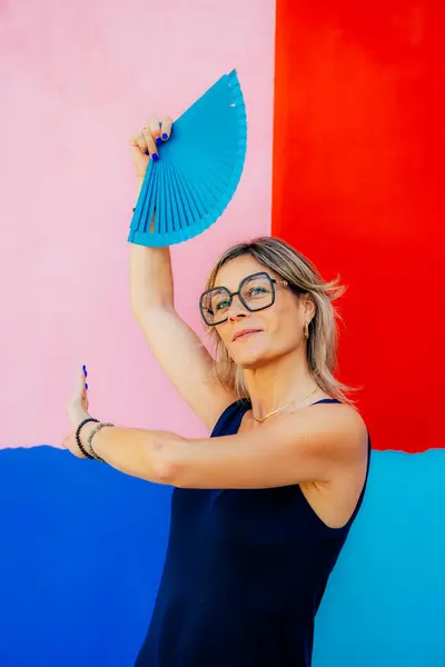 stock image A woman in a navy top strikes a playful pose with a blue fan against a colorful background, exuding confidence and joy.