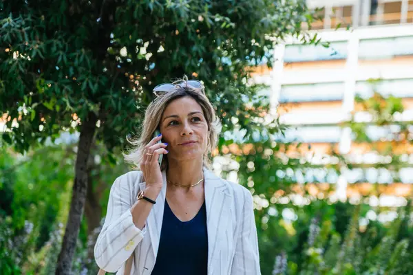 stock image A professional woman in a white blazer and navy top talks on her phone in an urban park.